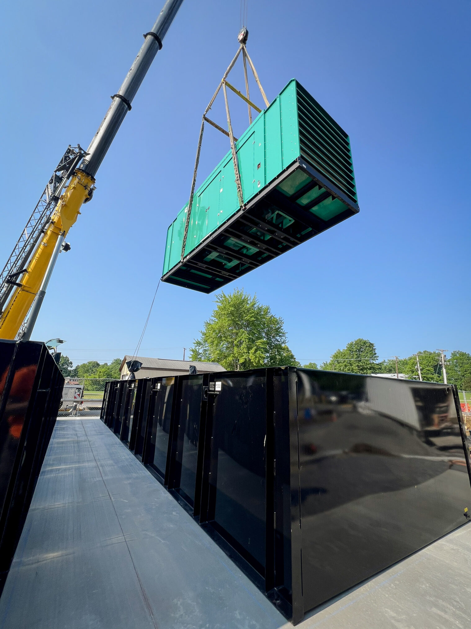 A large crane hoists a green industrial generator above black metal containers on a concrete surface, with a clear blue sky and green trees in the background.