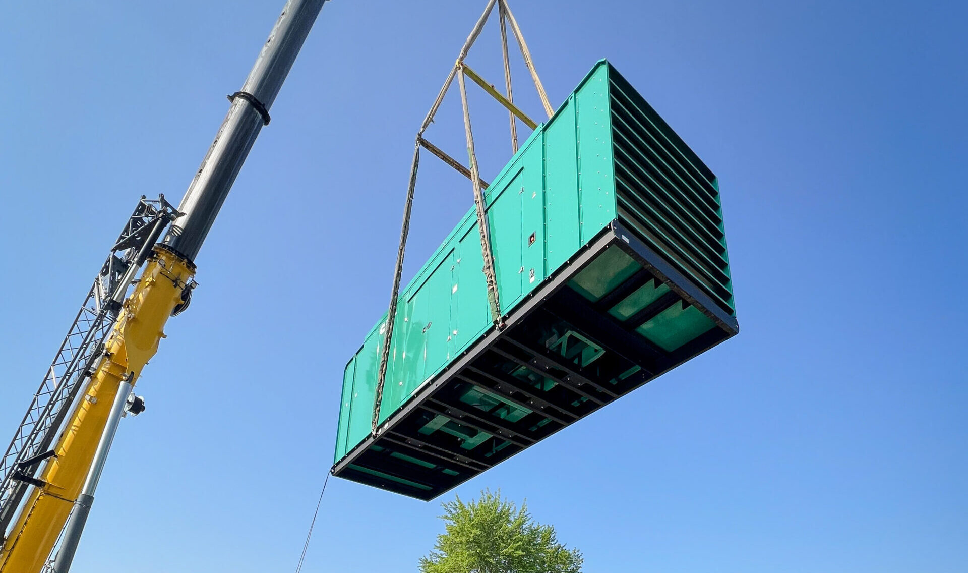 A large crane hoists a green industrial generator above black metal containers on a concrete surface, with a clear blue sky and green trees in the background.