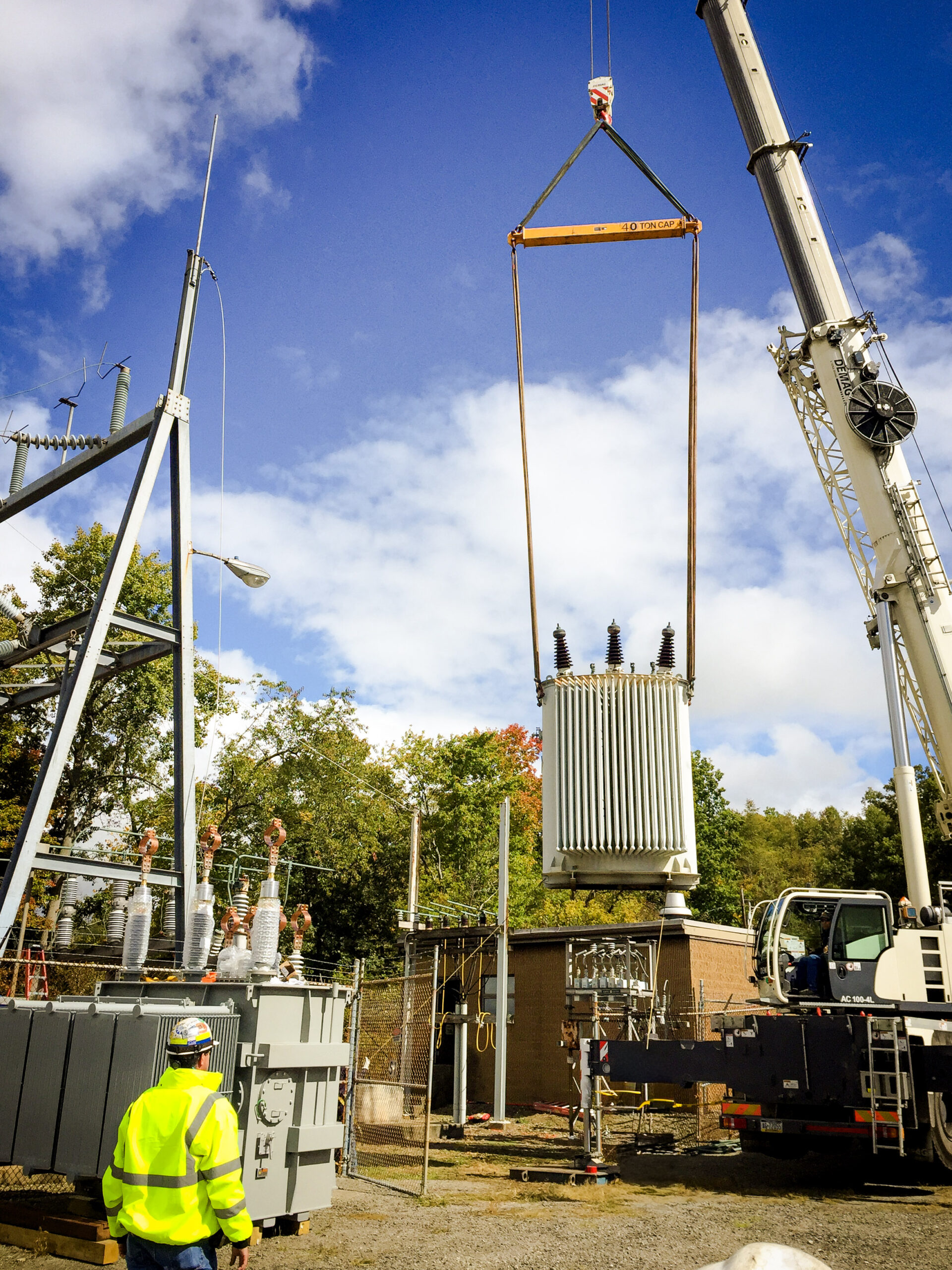 A large crane lifts a heavy electrical transformer at a power station, while a worker in a neon safety jacket and helmet observes from the ground. The sky is partly cloudy with trees in the background.