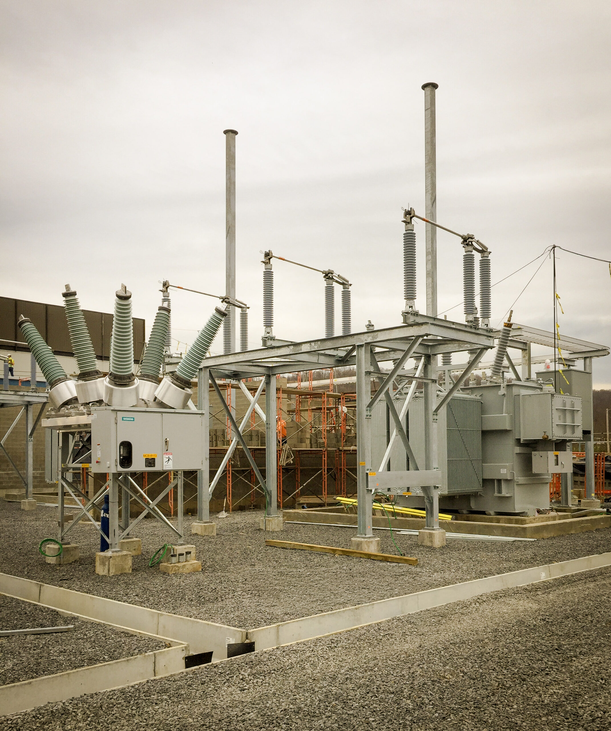 A power substation with various electrical equipment, including transformers, switches, and insulators, situated on a gravel lot under a cloudy sky. Scaffolding and construction materials are visible in the background.