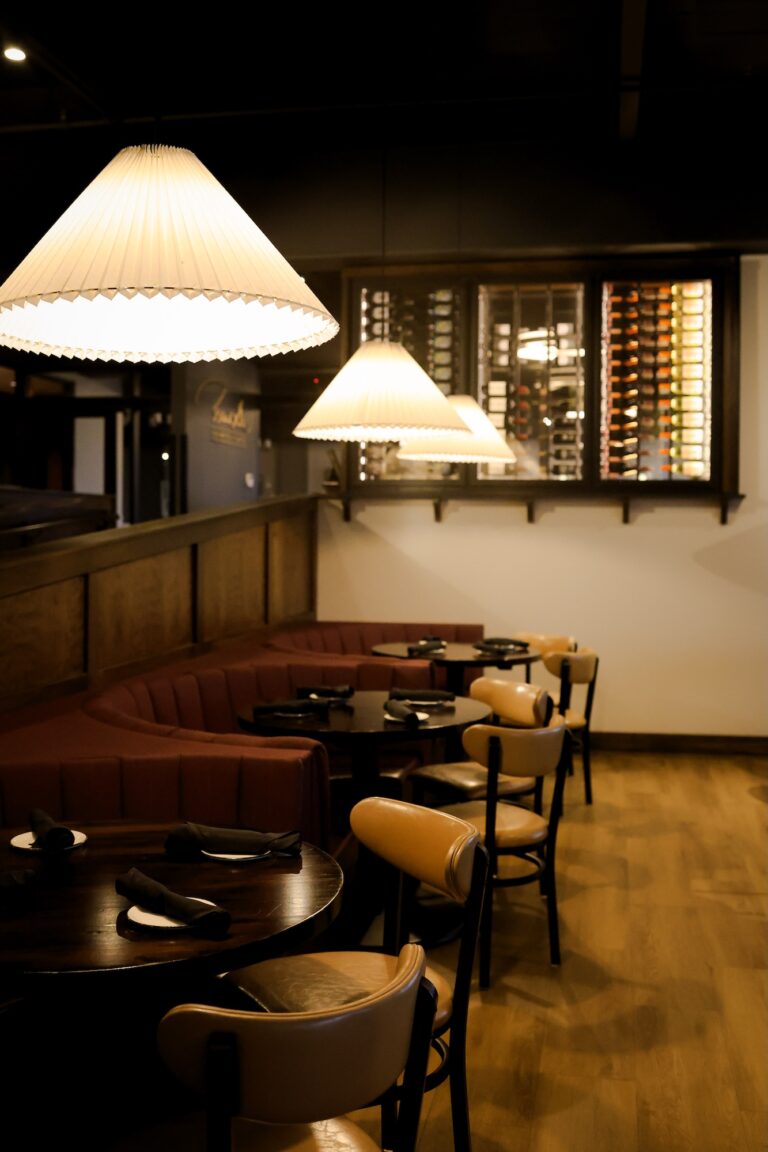 A warmly lit restaurant interior with round tables set for dining, cushioned chairs, a curved red booth, large hanging lamps, and a wine rack displayed on the back wall.
