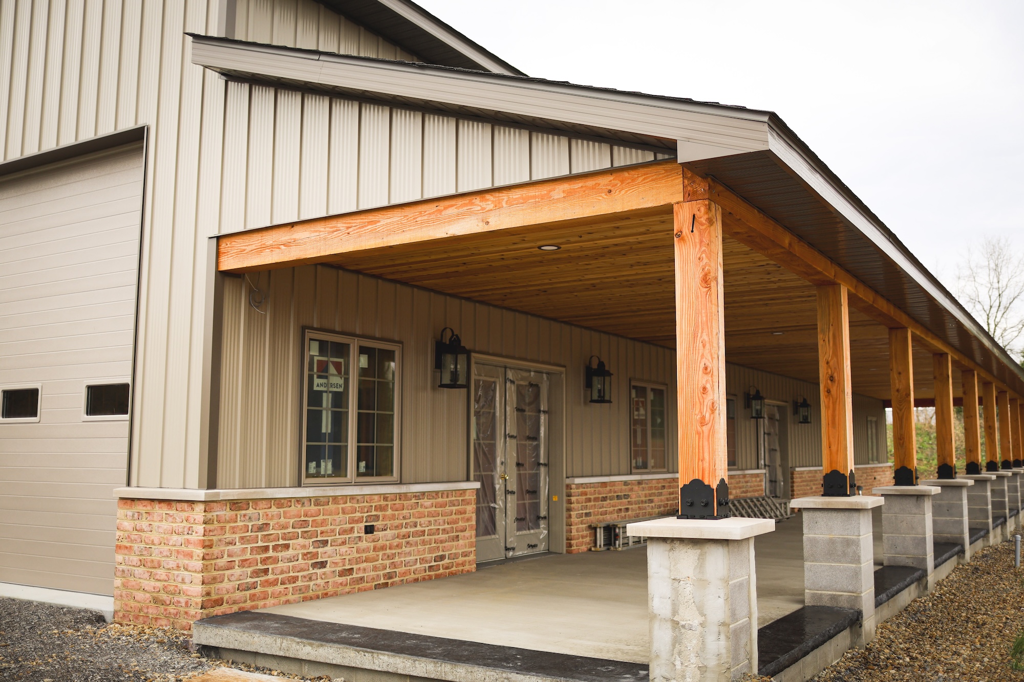 A modern building with tan metal siding, brick accents, a wooden porch roof supported by thick wooden columns, and a concrete walkway in front. Multiple windows and decorative lanterns are visible.