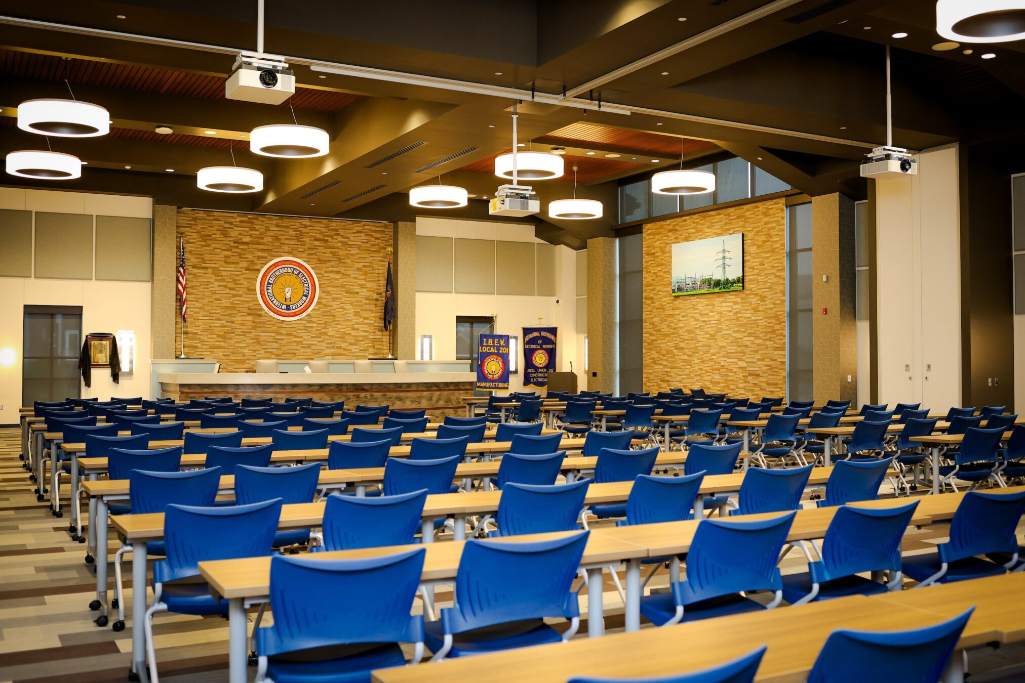 A large, modern conference room with rows of blue chairs facing a stage with a long desk, official seal, and banners. Circular lights hang from the ceiling, and a framed picture and American flag are on the back wall.