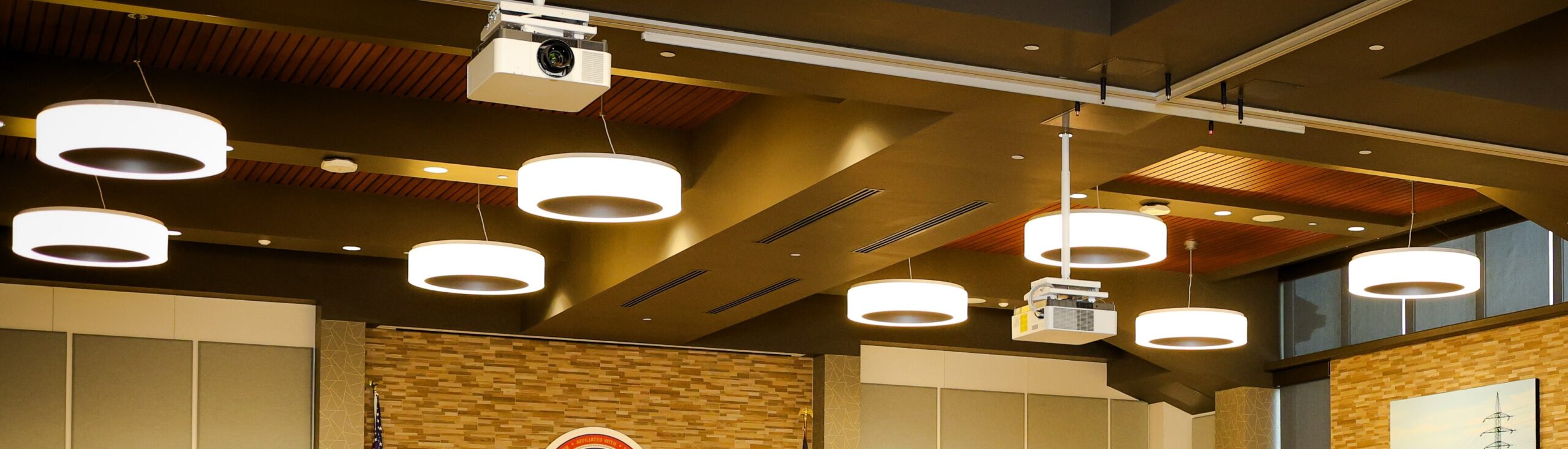 Ceiling of a modern room featuring circular pendant lights, two ceiling-mounted projectors, and sections of wood paneling with a brick accent wall and tall windows in the background.