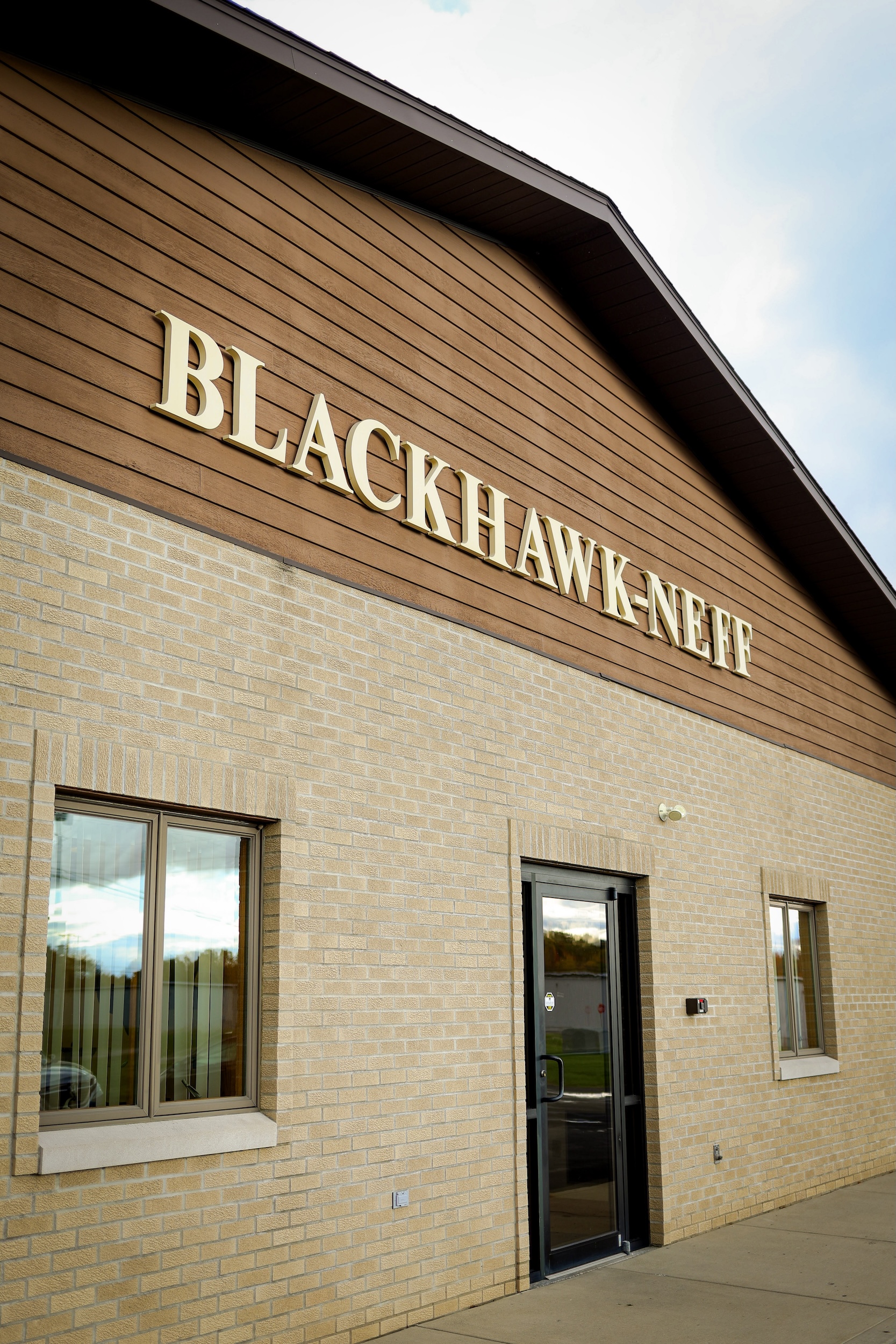 Exterior view of a tan brick building with a brown upper facade displaying large gold letters that spell BLACKHAWK-NEFF above a glass entrance door and two windows.