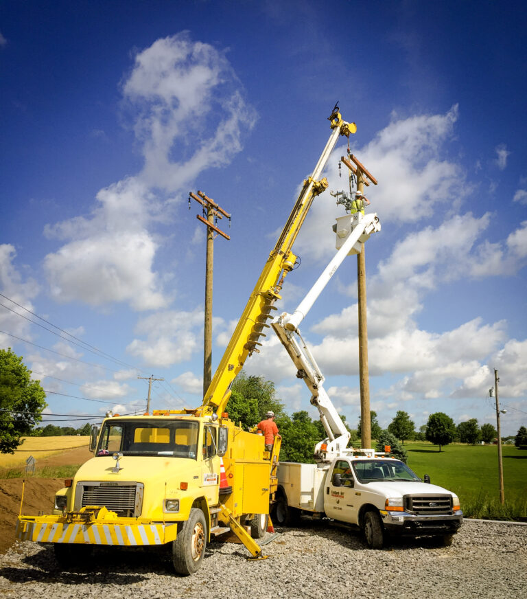 Two utility workers in bucket trucks repair power lines on tall poles under a bright blue sky with scattered clouds, surrounded by fields and trees.