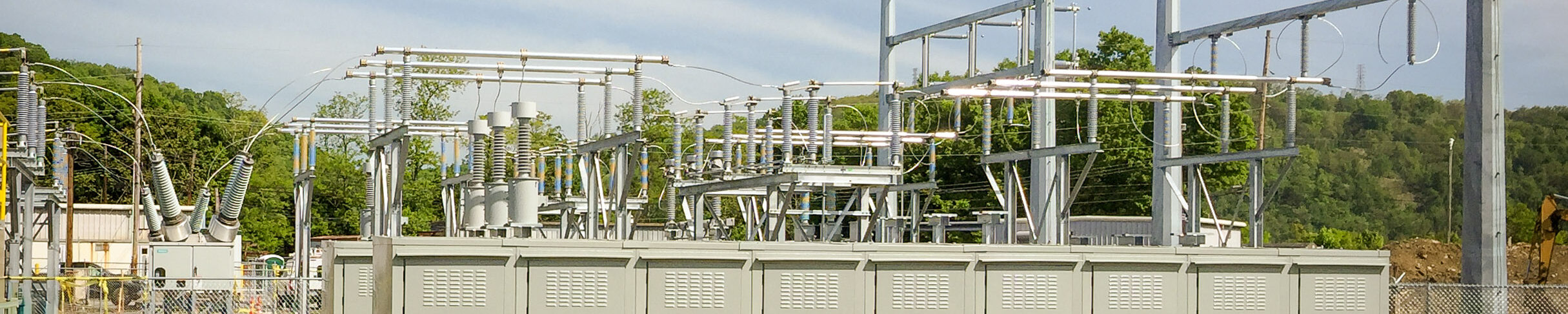 Outdoor electrical substation with metal structures, transformers, and power lines, surrounded by a metal fence, set against a backdrop of green trees and clear sky.