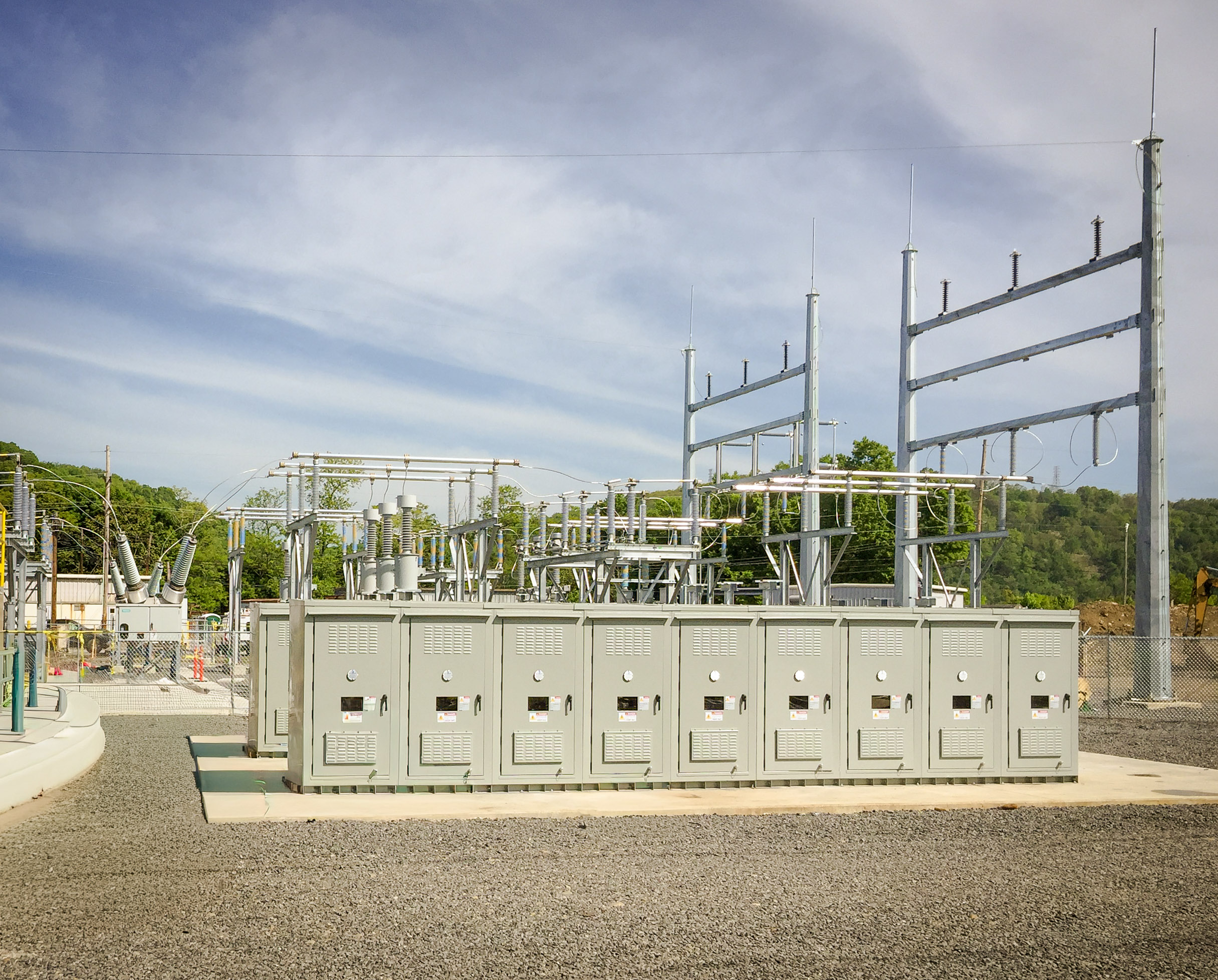 An outdoor electrical substation with metal structures, power lines, and gray control panels, set on a gravel-covered ground with trees and hills visible in the background under a partly cloudy sky.