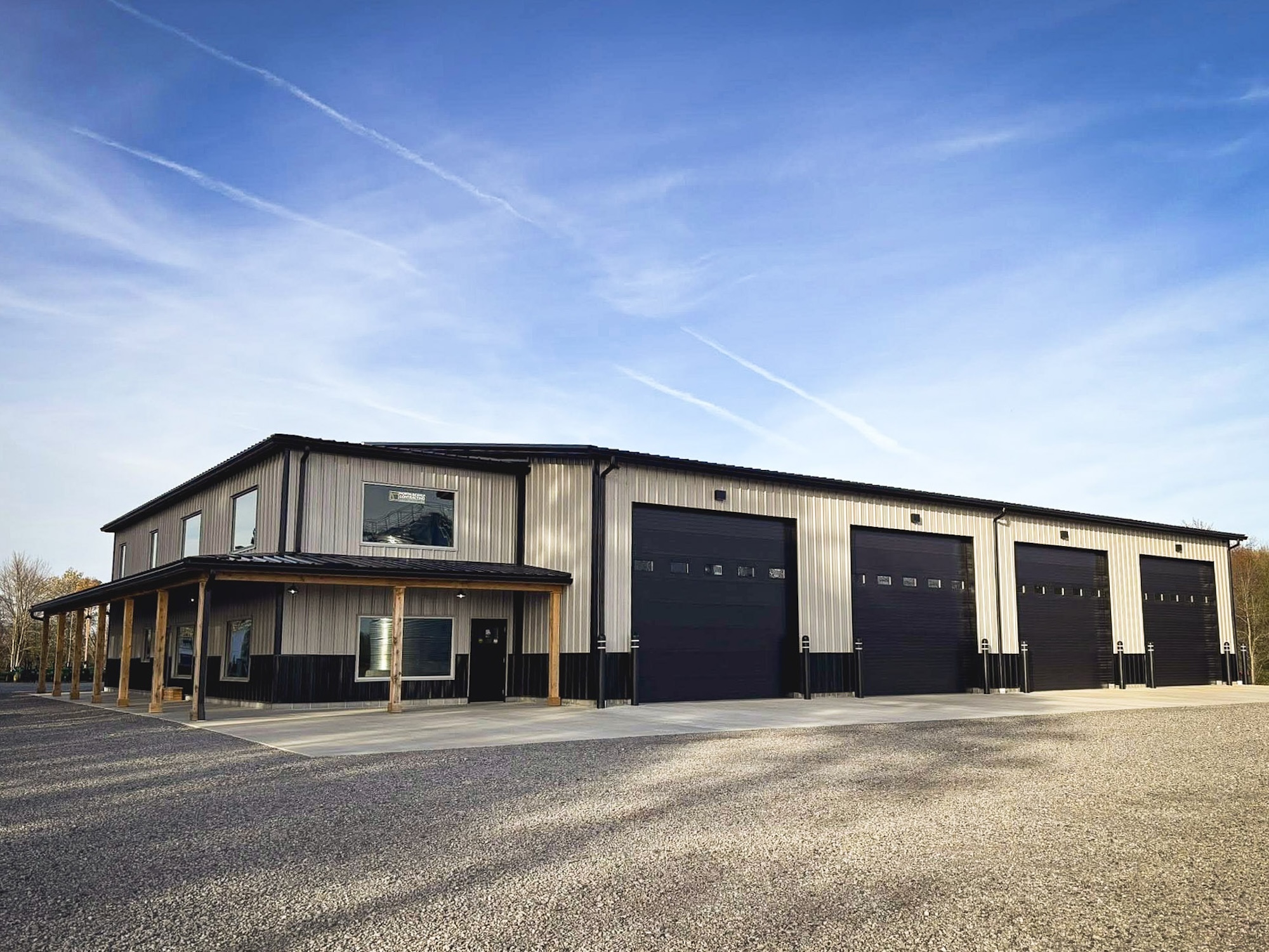 A large, modern metal building with four tall garage doors and a covered entrance sits on a gravel lot under a clear blue sky with contrails.