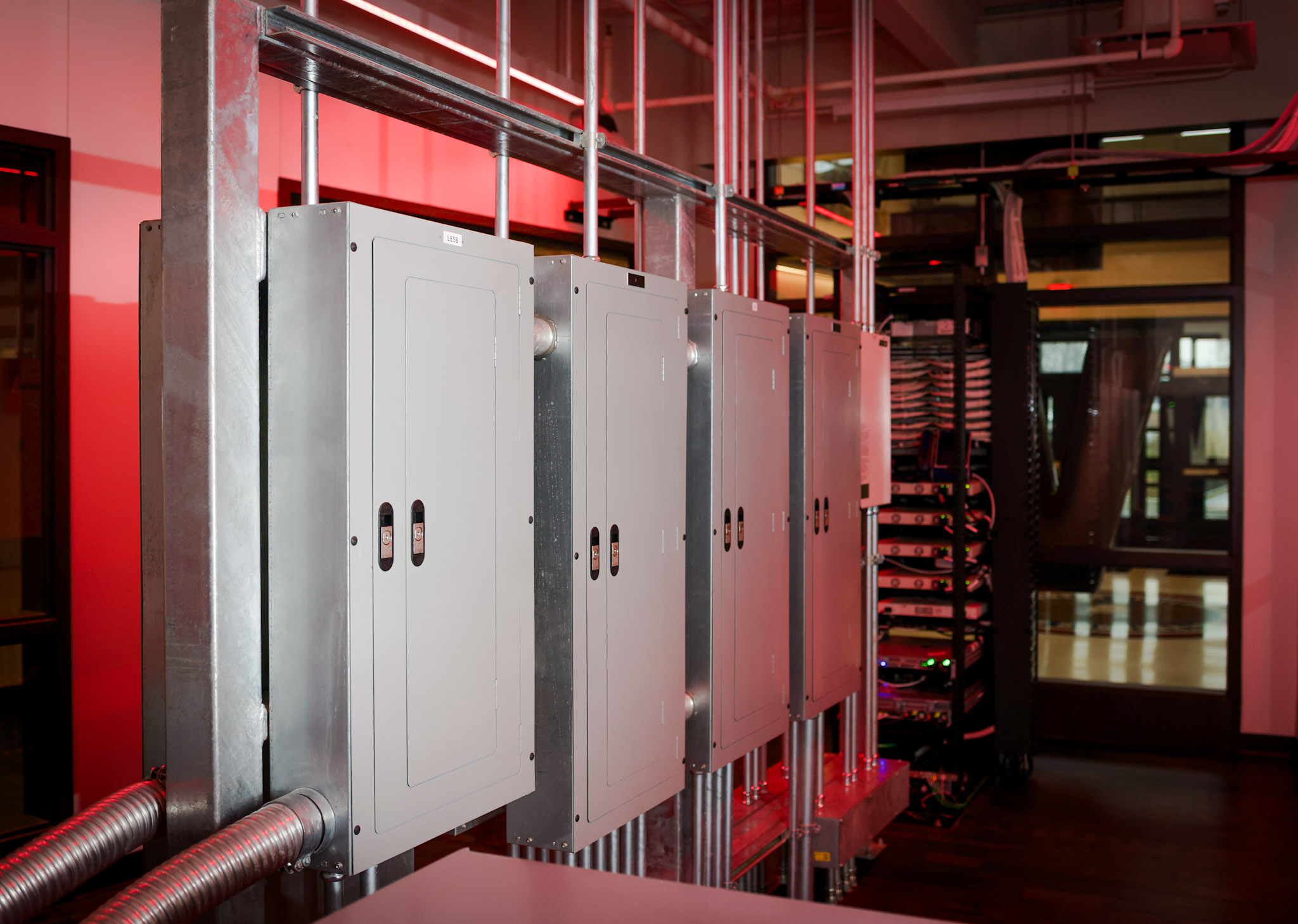 A row of metal electrical panels is mounted on a wall with exposed pipes, with a server rack and red ambient lighting in a modern industrial room.
