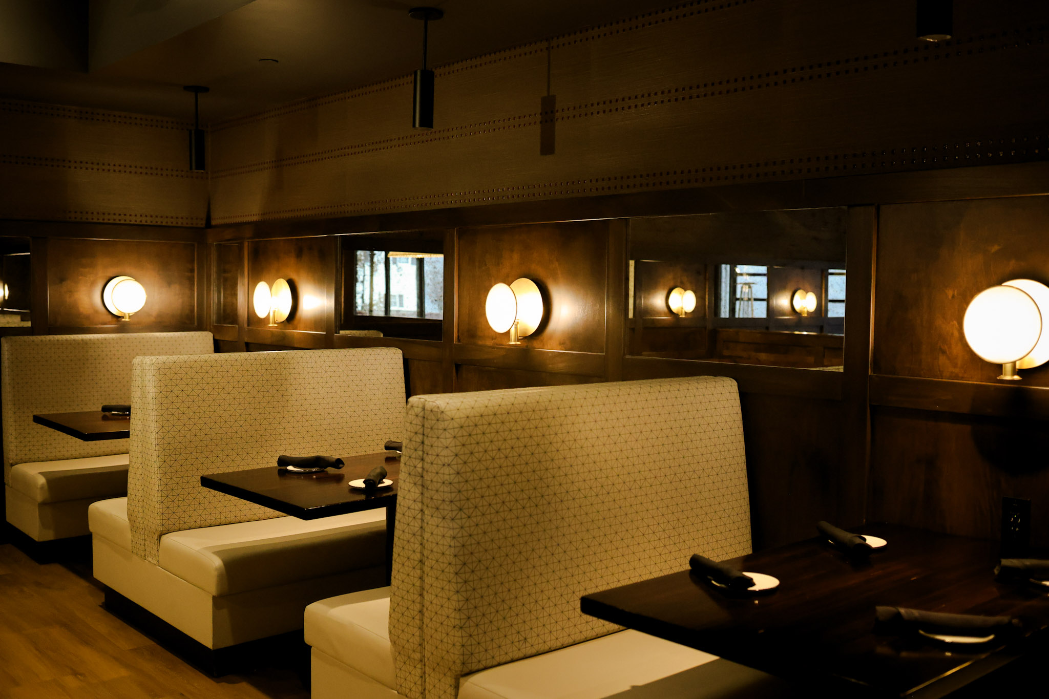 A row of empty restaurant booths with cushioned seats and dark wooden tables, set with napkins and utensils, softly lit by round wall-mounted lamps above mirrors. The atmosphere is warm and inviting.