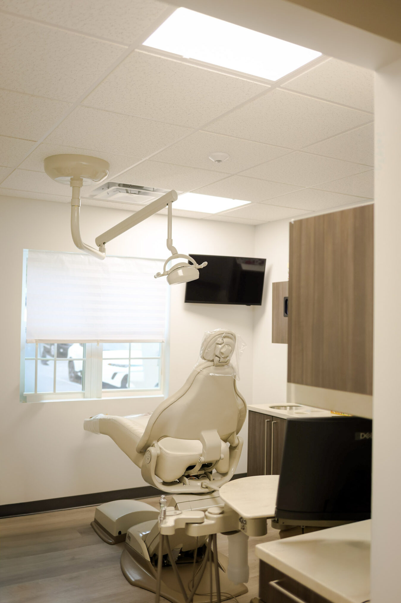 A modern dental office with a beige dental chair, overhead light, wall-mounted TV, cabinets, a window with blinds, and various dental equipment arranged neatly in a clean, well-lit room.