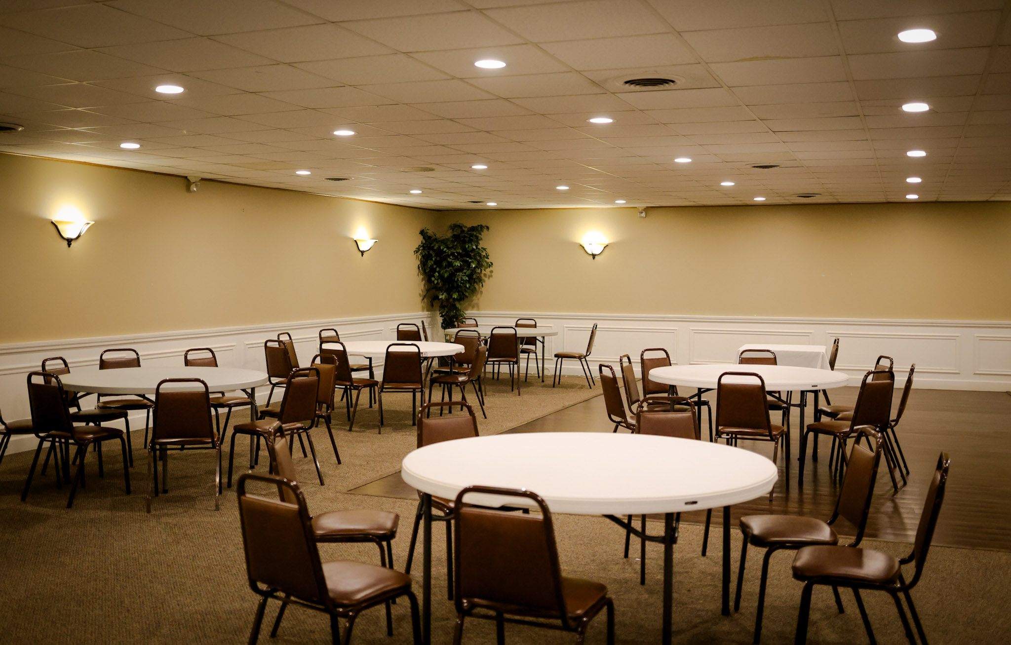 A spacious, empty banquet room with round white tables and brown chairs arranged in small groups. Beige walls, white trim, and wall-mounted lights create a warm atmosphere; a potted plant is in the corner.