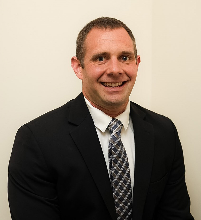 A man in a black suit, white shirt, and patterned tie is smiling while sitting against a plain light-colored wall.