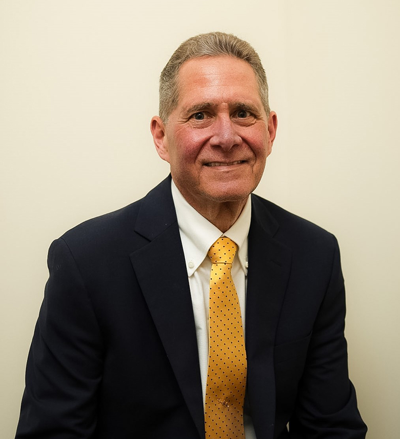 A man in a dark suit, white shirt, and yellow polka dot tie sits smiling against a plain, light-colored background.
