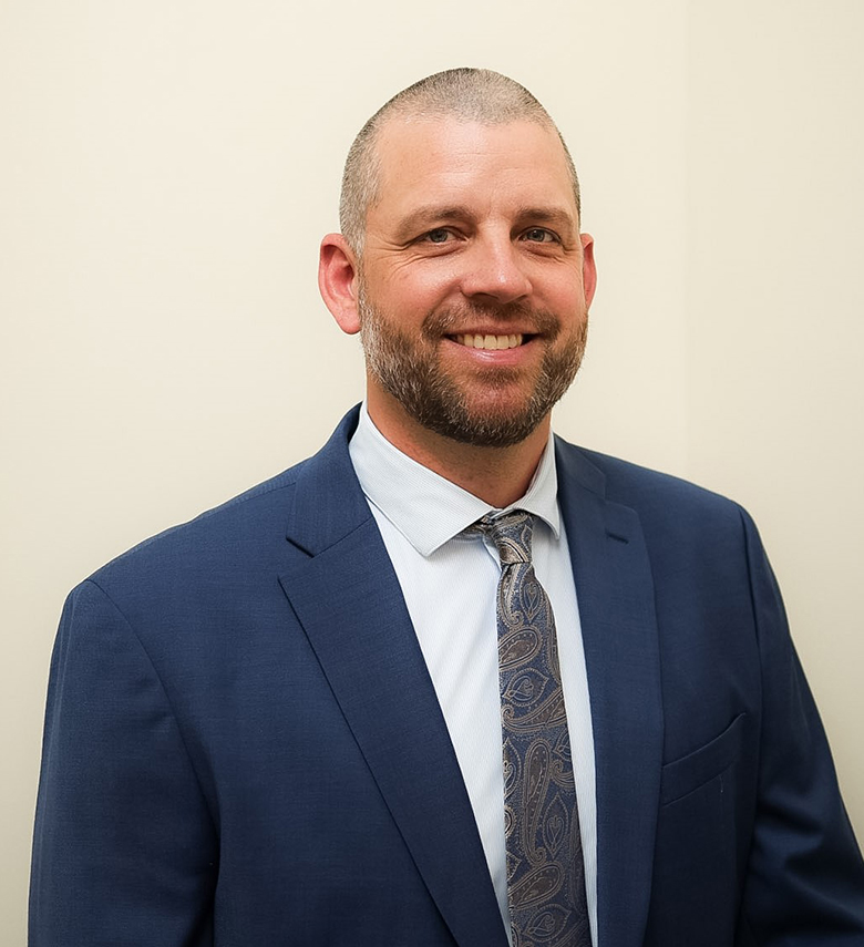 A man with a short beard and closely-cropped hair, wearing a blue suit, white shirt, and patterned tie, smiles while standing against a plain light-colored background.