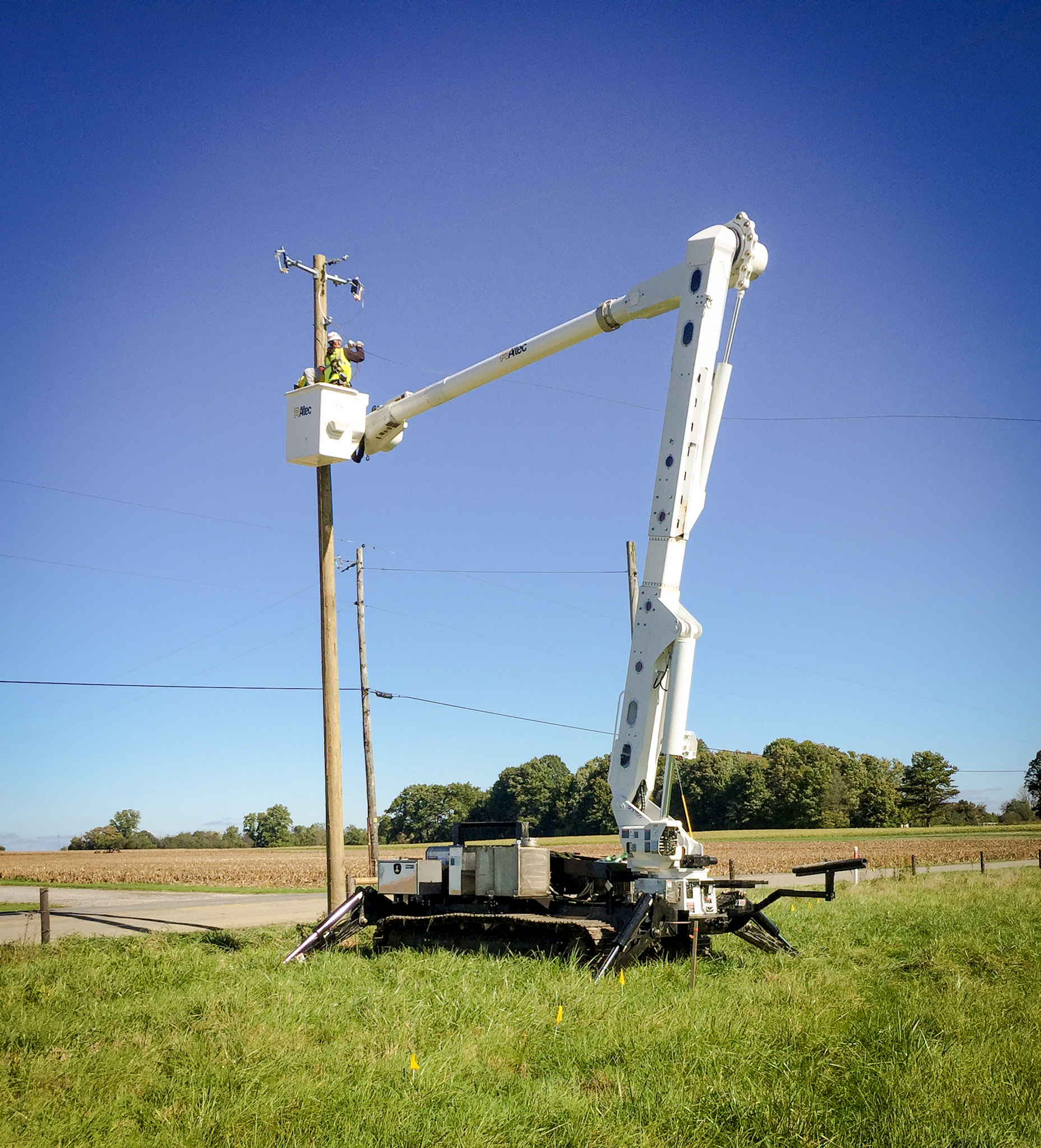 A utility worker in a raised bucket truck repairs power lines on a wooden pole in a rural field under a clear blue sky. The vehicle’s stabilizing legs are extended for support on the grass.