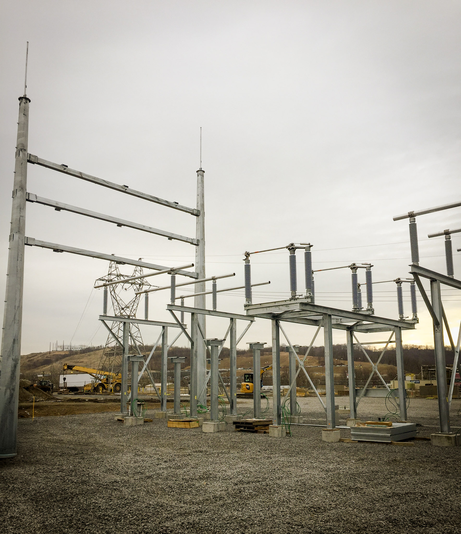 A construction site with metal frameworks being assembled for an electrical substation, with cranes and trucks in the background under an overcast sky.