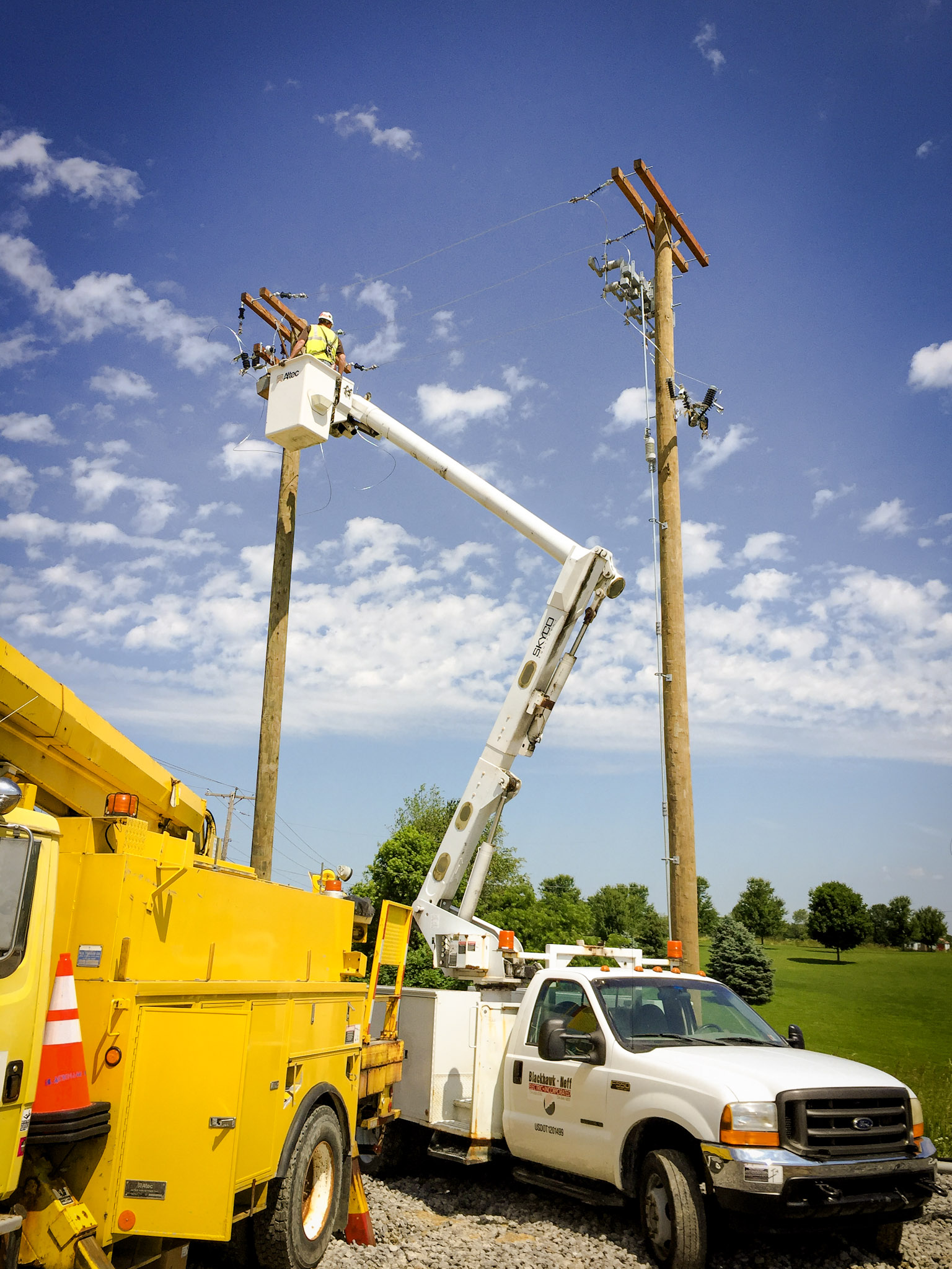 A utility worker in a raised bucket truck repairs power lines on a wooden pole under a blue sky. Nearby, another yellow utility truck is parked on gravel beside a grassy field with trees in the background.