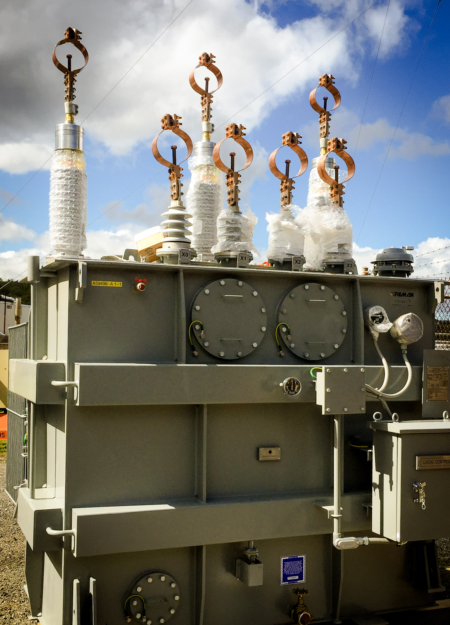 Large electrical transformer outdoors with multiple high-voltage bushings wrapped in protective plastic at the top, set against a partly cloudy sky and surrounded by a metal fence.