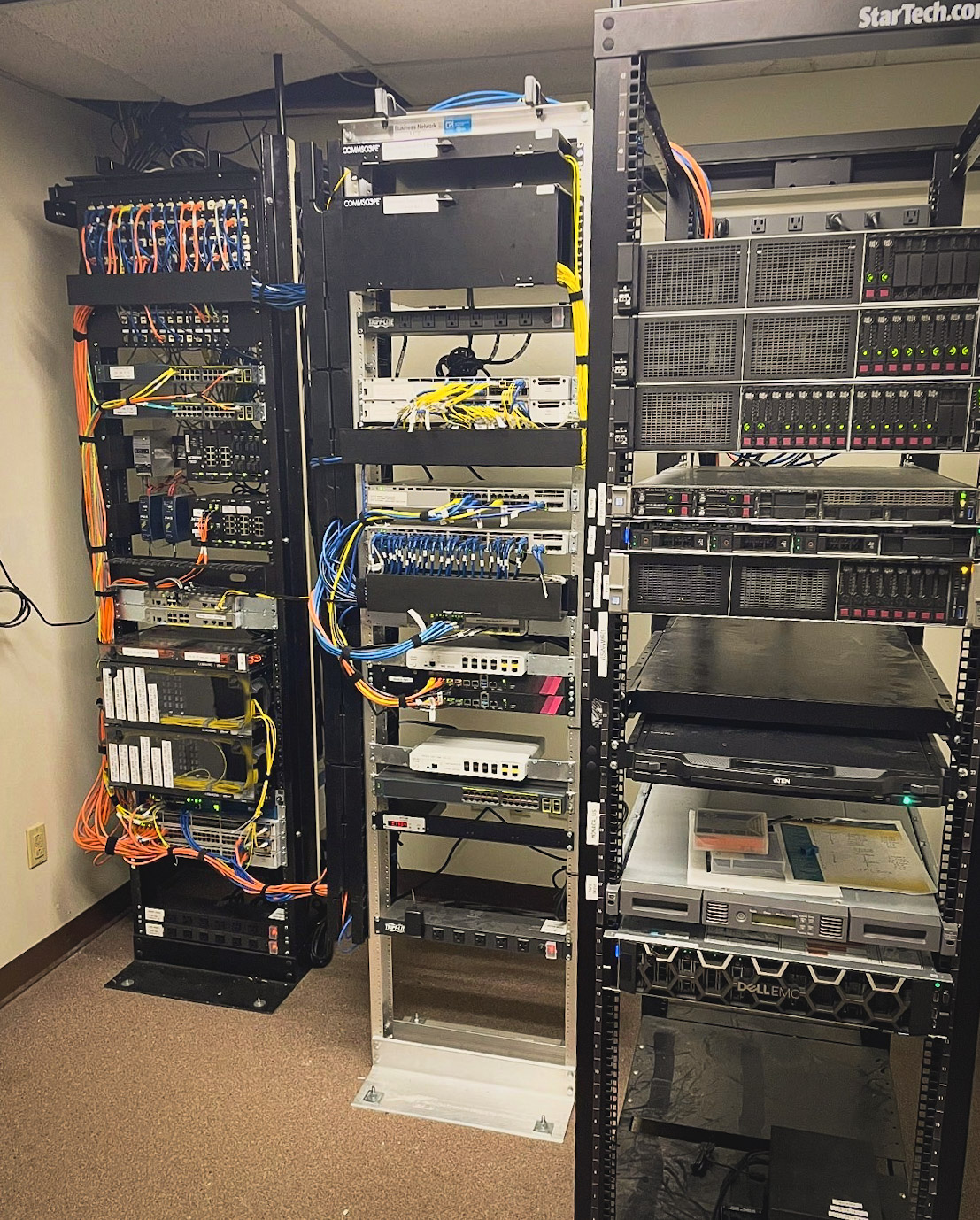 A server room with three racks filled with network switches, servers, and various cables organized and connected to the equipment, all standing on a carpeted floor against a beige wall.