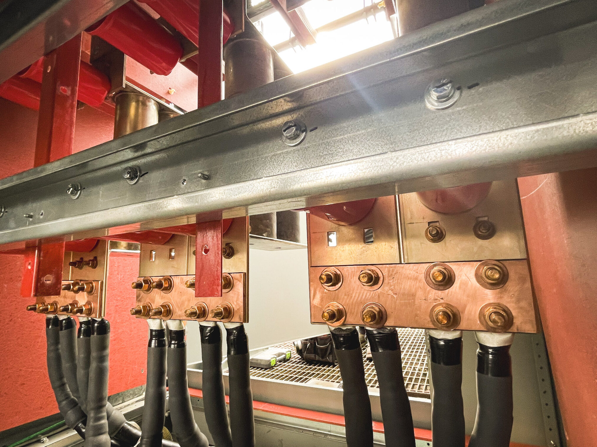 Close-up view of industrial electrical busbars and copper terminals connected by thick black cables, with metal bolts securing the connections inside a facility. Red and metallic structural elements are visible in the background.