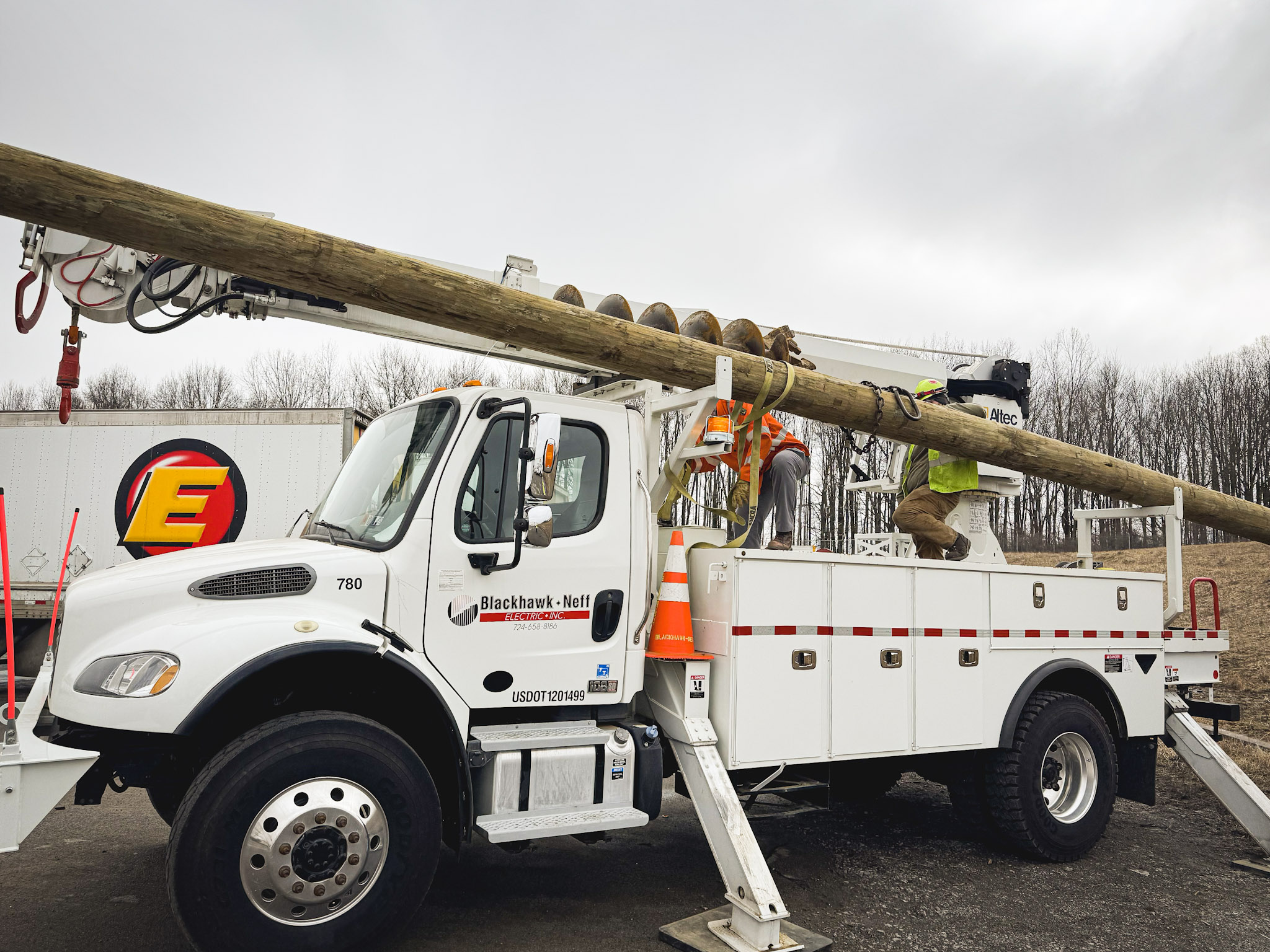 Two utility workers in safety gear stand on a platform attached to a white utility truck, positioning a tall wooden pole with equipment on an overcast day. Leafless trees and a truck are visible in the background.