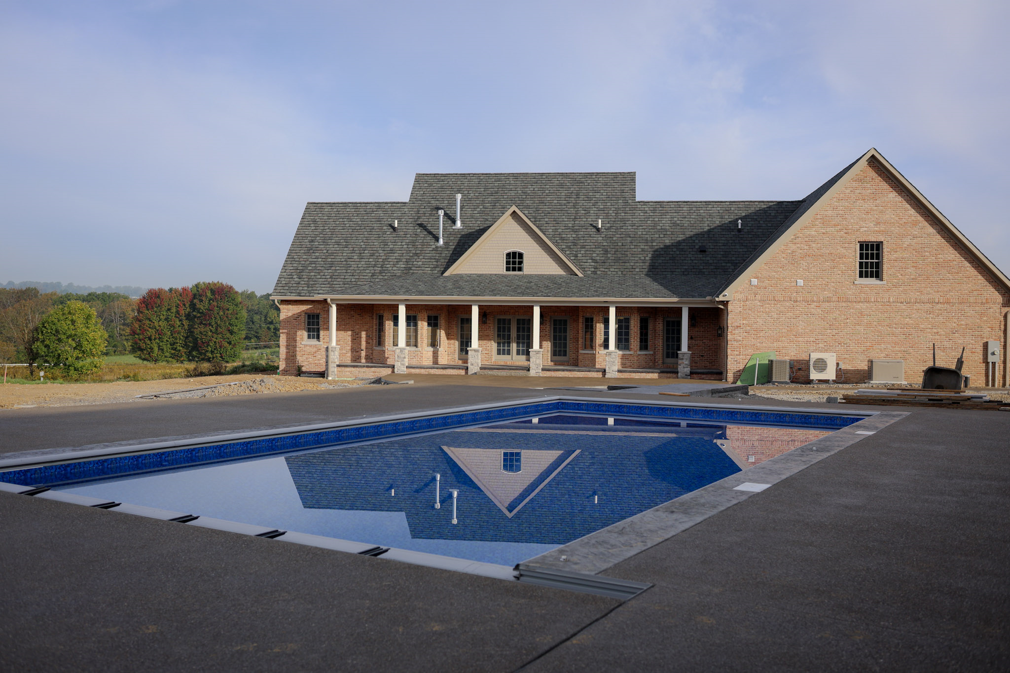 A brick house with a gray roof stands behind a rectangular in-ground swimming pool on a concrete patio, with trees and blue sky reflected in the pools water.