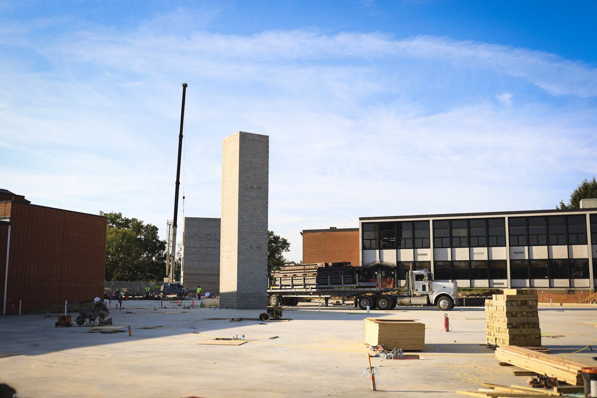 A construction site with a tall, rectangular concrete structure, a crane, a truck, building materials, and workers near modern and brick buildings under a blue sky.