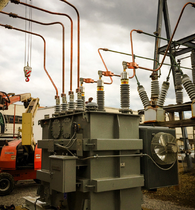 A large industrial electrical transformer with several thick copper wires and insulators is installed outdoors. Nearby, a crane and other equipment are visible under a cloudy sky.