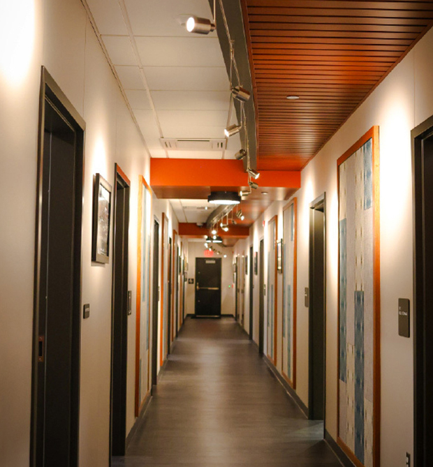 A well-lit hallway with several closed doors on both sides, light-colored walls, framed art pieces, and a dark floor, leading to a black door at the end. The ceiling features exposed fixtures and wooden paneling.