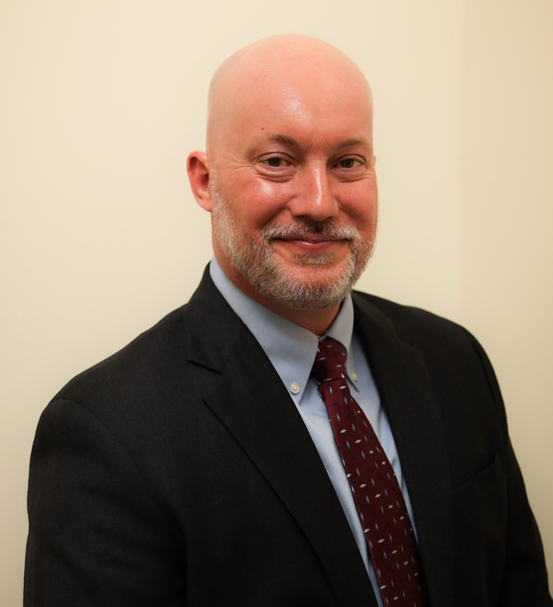 A bald man with a trimmed gray beard wearing a dark suit, light blue shirt, and maroon patterned tie, smiling and posing in front of a plain light-colored background.
