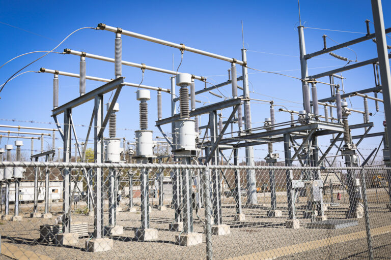 An electrical substation with metal structures, insulators, and power lines, surrounded by a chain-link fence, under a clear blue sky.