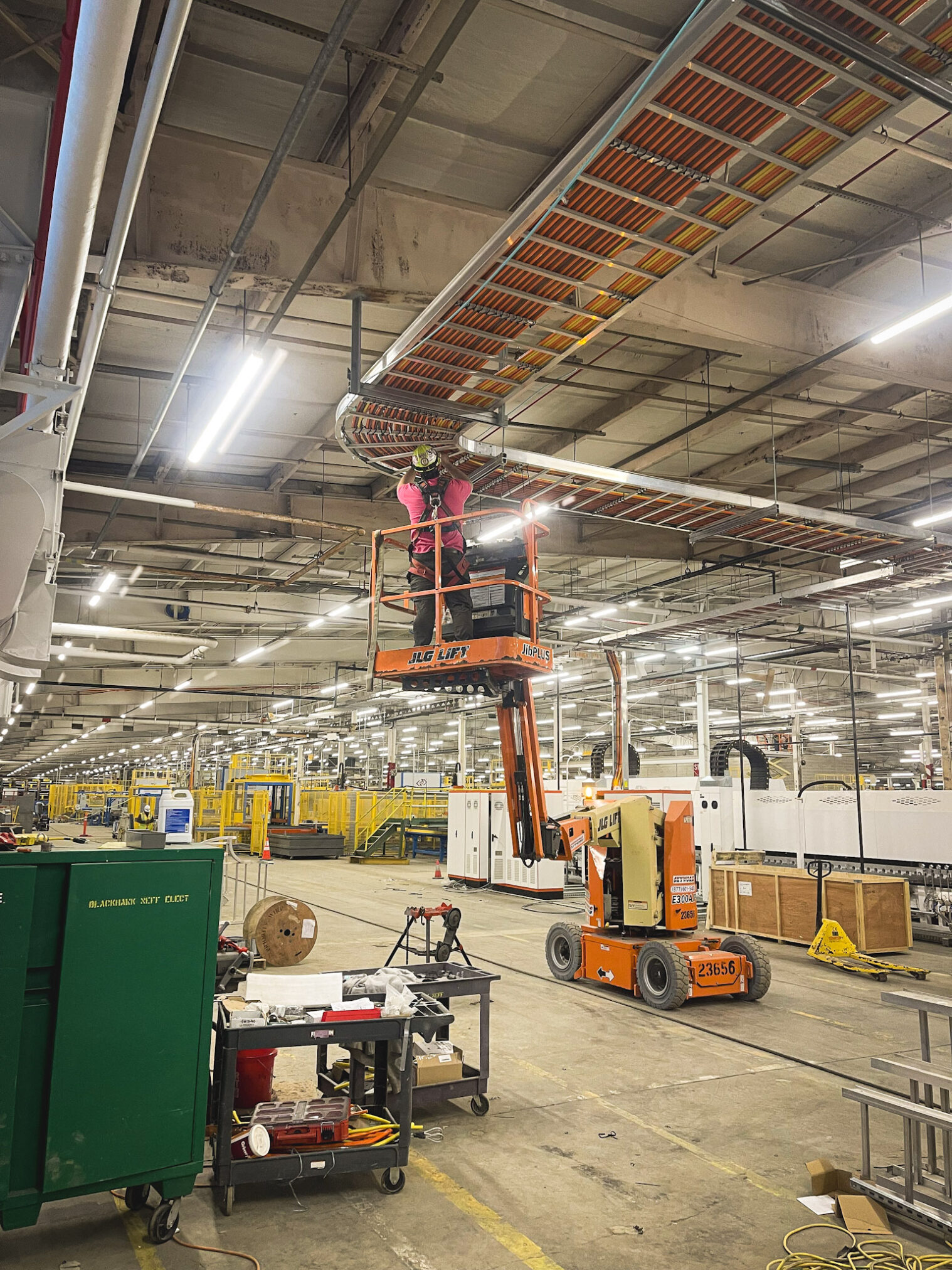 Two workers in safety gear stand on an elevated lift, working near overhead pipes and cables inside a large industrial warehouse with tools, equipment, and machinery visible below.