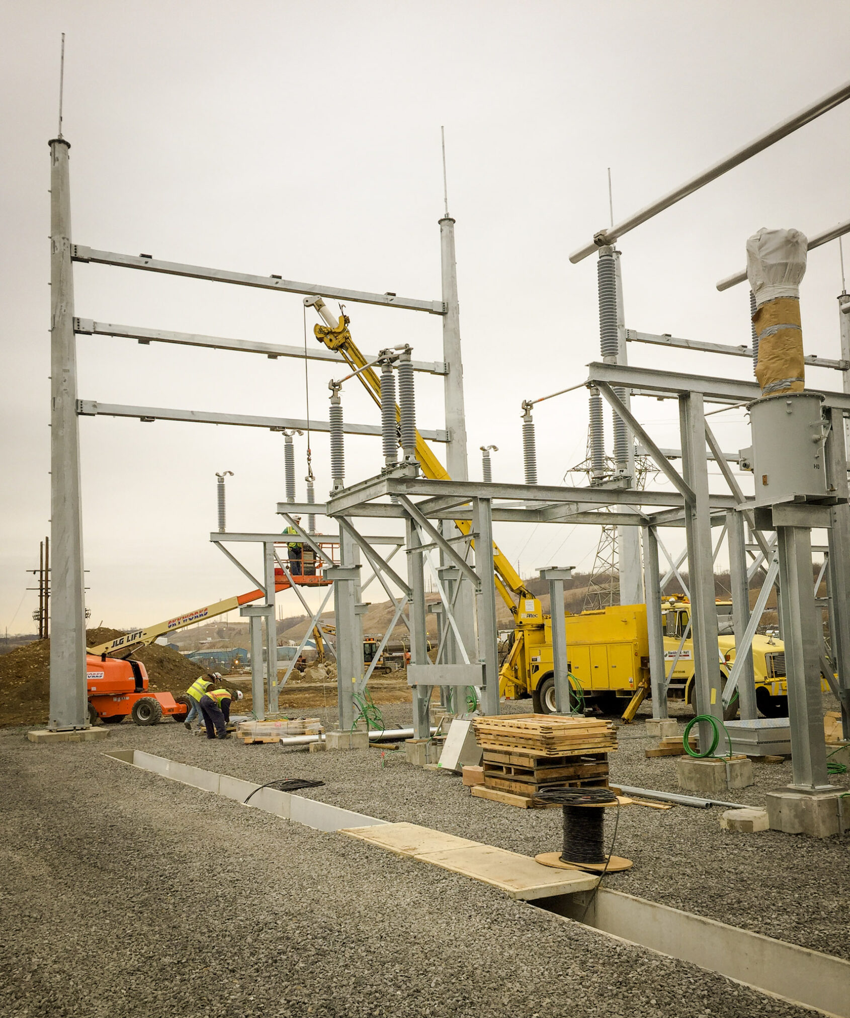 Construction workers install electrical equipment at a substation. Yellow cranes and trucks are present, along with metal structures and insulators.
