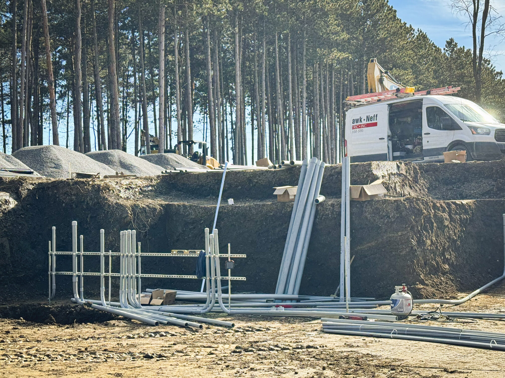 Construction site with exposed soil, stacked metal pipes, and gravel piles. A white work van and equipment are present, with tall pine trees in the background under a clear blue sky.