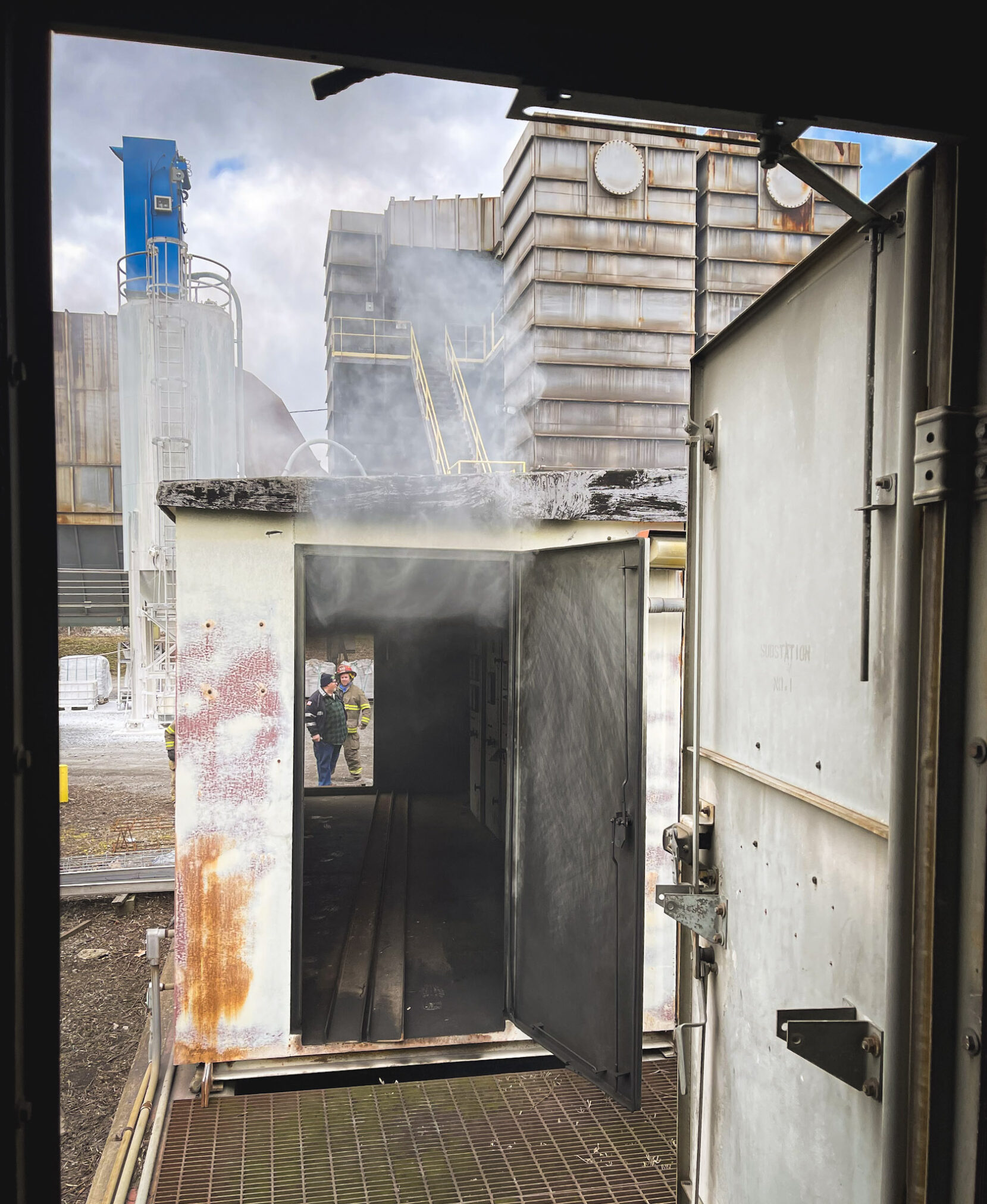 A metal shipping container with smoke rising from its roof stands open, revealing a dark interior. Industrial buildings and a silo are visible in the background, with two people in safety gear standing outside.
